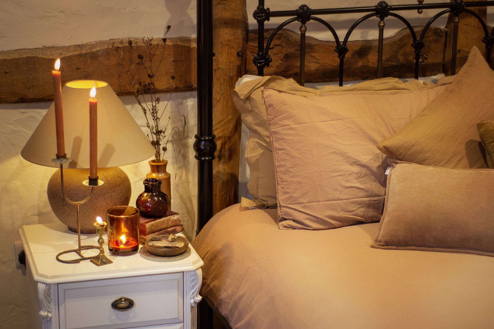 Candlelit bedside table beside an iron bed made up in warm terracotta organic cotton bedding under old oak beams