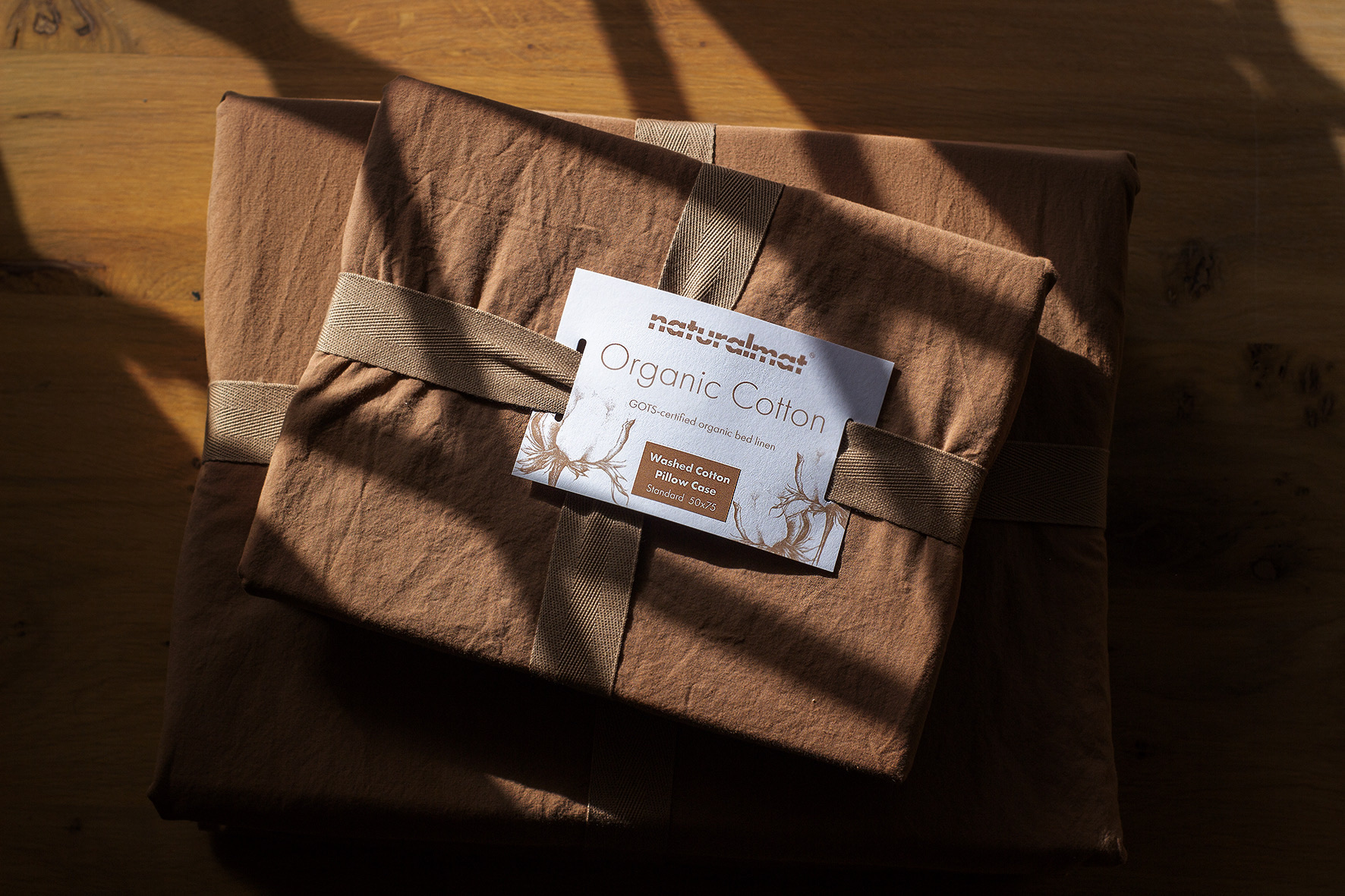 Naturalmat washed organic cotton pillowcase in terracotta tied with cotton tape on an oak table in low sunlight