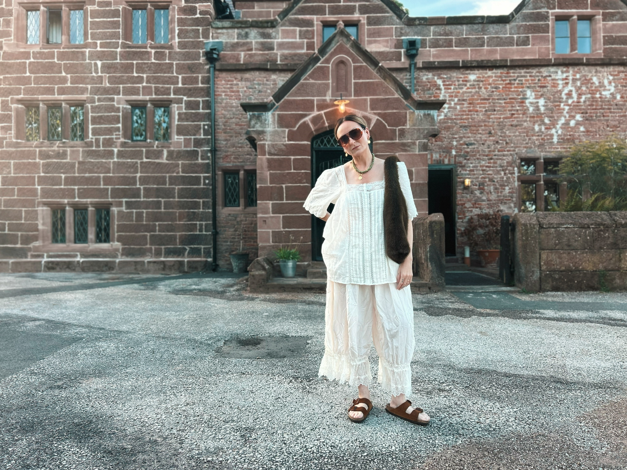 Susannah in a white lace blouse and bloomers standing in front of the sandstone manor house