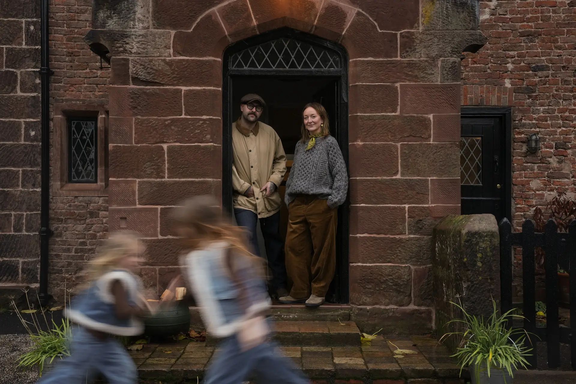 Susannah and Carl standing in the stone doorway of the manor while their two daughters run past
